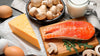 Erilaisia D-vitamiinin lähteitä pöydällä, kuten kalaa, juustoa, sieniä ja maitoa.